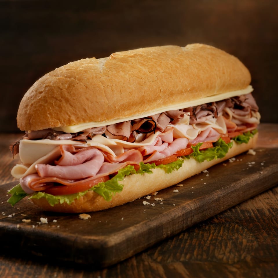 A large Mancino's Grinder sandwich, filled with ham, roast beef, cheese, lettuce, and tomato, on a wooden board.