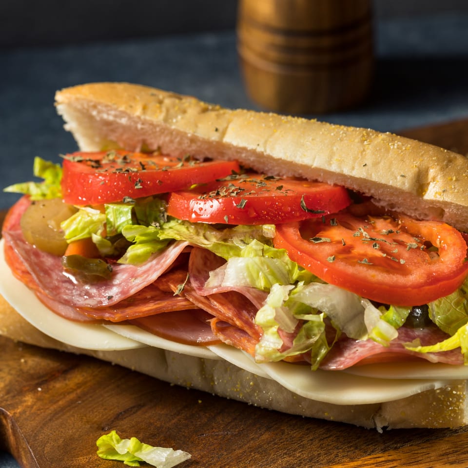 A Mancino's Grinder sandwich filled with salami, cheese, lettuce, tomatoes, and pickles on a wooden board.