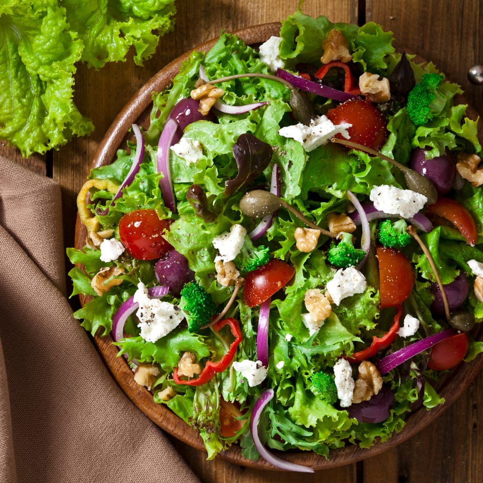 A vibrant salad with lettuce, tomatoes, red onion, feta, walnuts, and capers in a wooden bowl.