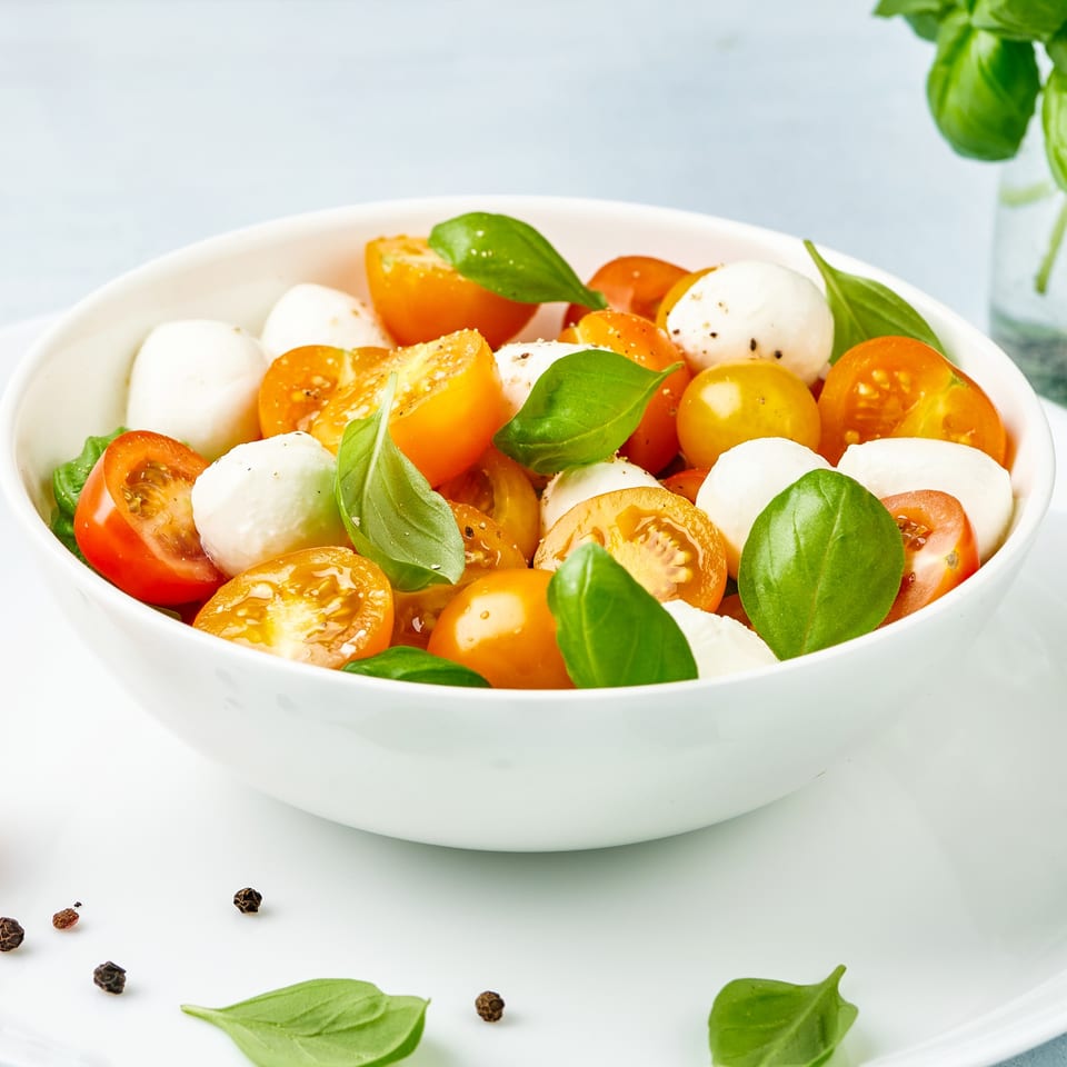 A white bowl filled with fresh mozzarella balls, halved yellow and red cherry tomatoes, and basil leaves.